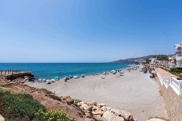 A coastal beach with a rocky shoreline, a promenade with buildings, and a clear blue sky. - Toboso 1 (Nerja)