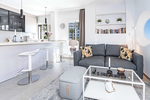Una sala moderna con un sofá gris, una mesa de centro de cristal y una cocina con taburetes blancos.