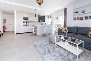 A modern living room with a grey sofa, a coffee table, and a kitchen area with white cabinets and a bar stool. - Toboso 1 (Nerja)