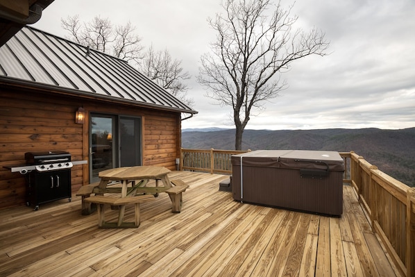 A wooden deck with a grill and hot tub, overlooking a mountainous landscape.