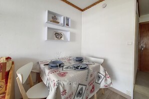 Uma sala de jantar com uma mesa posta para quatro pessoas, cadeiras brancas e toalha de mesa branca com padrão. Há prateleiras acima da mesa com objetos decorativos.