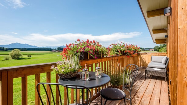 Una terraza de madera con una mesa y sillas, un espacio de descanso acolchado y macetas con flores.