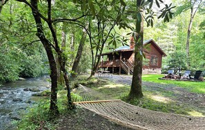 Una cabaña de madera con balcón, rodeada de árboles y un arroyo.