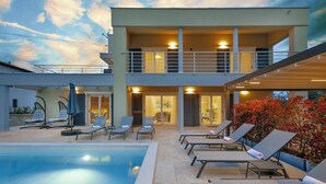 A modern two-story building with a swimming pool, lounge chairs, and a covered patio area.