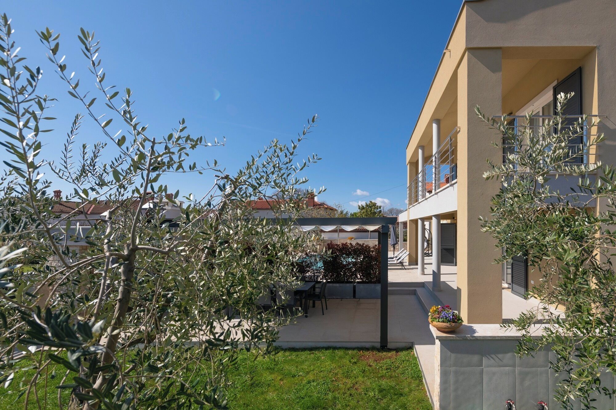Ein modernes Gebäude mit überdachter Außenbestuhlung, einem Pool und einem Garten mit Olivenbäumen.