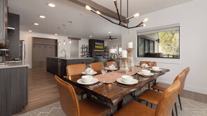 A modern dining area with a wooden table set for six, surrounded by leather chairs. A kitchen with dark cabinetry and a built-in island is visible in the background. - Beaverhead Village Haven | Hot Tub • Walk to Big Sky (Big Sky)
