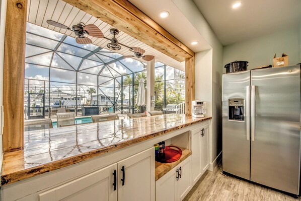 A kitchen with a large window overlooking a pool area, a stainless steel refrigerator, and a wooden ceiling.