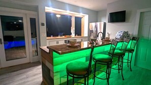 A modern kitchen with a bar area, green lighting, and a TV mounted on the wall.