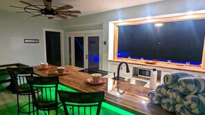 A modern kitchen with a wooden island, black chairs, a ceiling fan, and a large window.