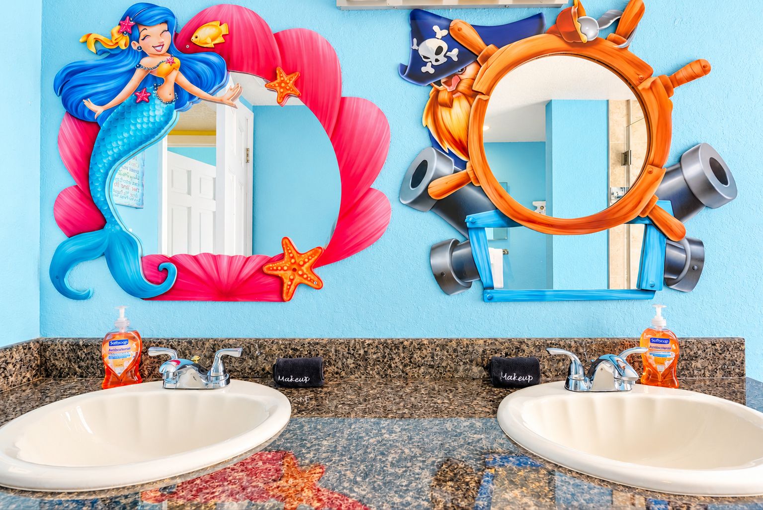 Une salle de bain avec deux lavabos, un miroir et des éléments décoratifs, notamment des œuvres d'art murales sur le thème des sirènes et des pirates.