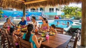A family dining at an outdoor table with a pool and a thatched roof in the background. - Beach House, Waterfront, Sombrero Beach, Florida Keys Rental, Private Pool, Dock (Marathon)