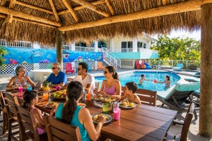 A family dining at an outdoor table with a pool and a thatched roof in the background.