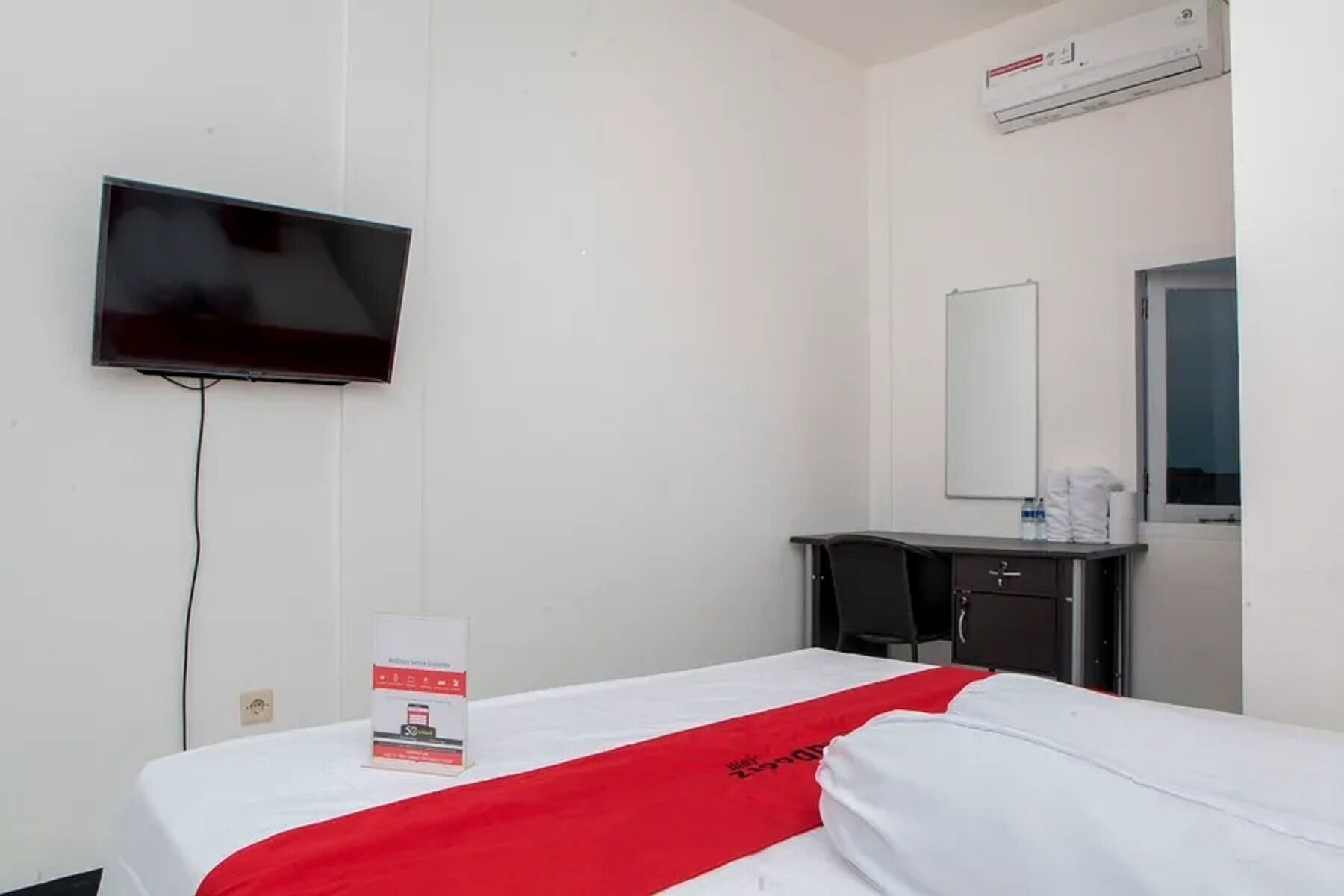 A hotel room with a bed, a TV mounted on the wall, a small desk with a mirror, and an air conditioning unit.