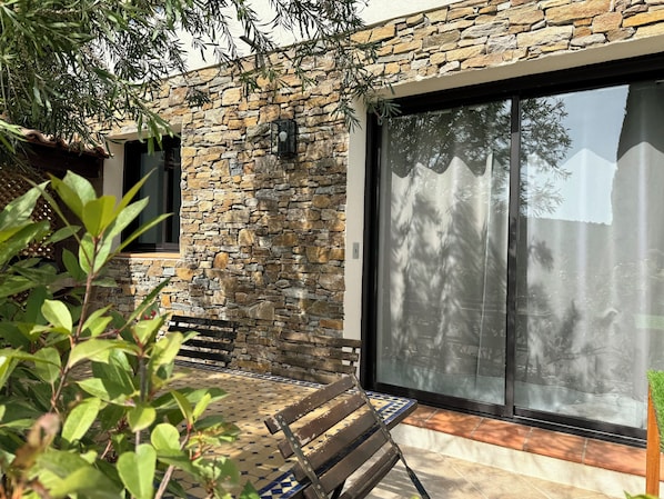 A stone exterior wall with a large sliding glass door, a wooden bench, and a tiled patio area.