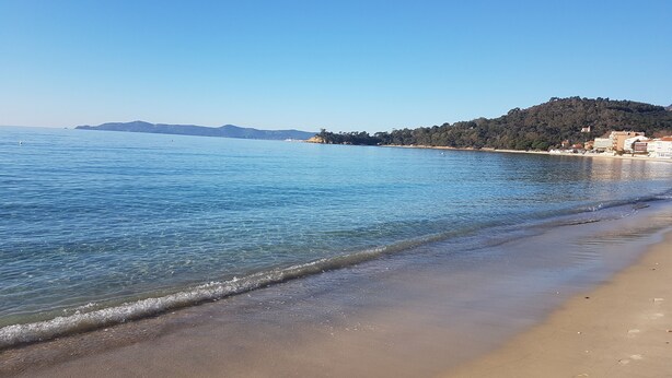 A beach with clear water, a sandy shore, and buildings on a hillside in the background.