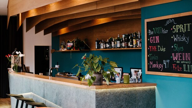 Zona de bar con un menú en pizarra que enumera bebidas, una planta en maceta y estantes con botellas.