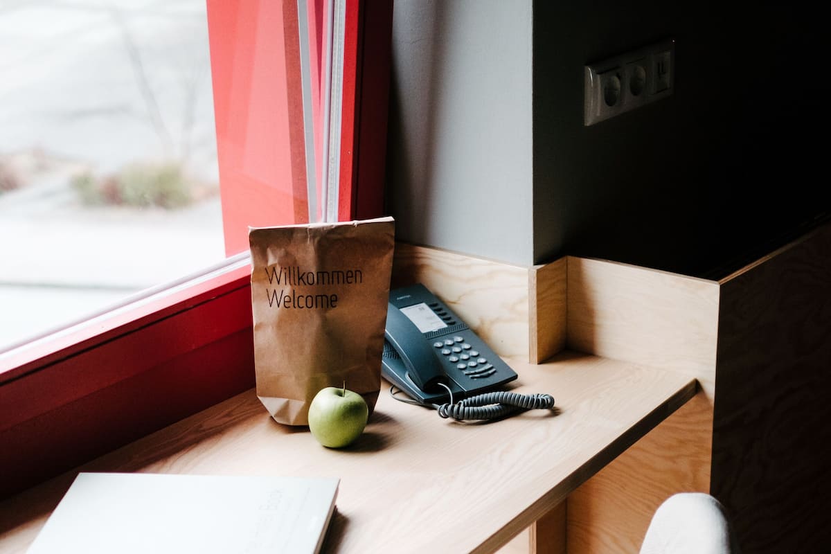 A wooden desk with a black chair, a book titled "The Hotel Book," a welcome bag, and a telephone.