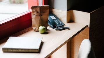 A wooden desk with a black chair, a book titled "The Hotel Book," a welcome bag, and a telephone.
