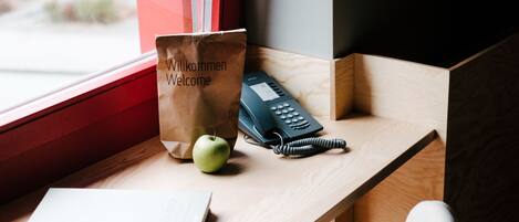 A wooden desk with a black chair, a book titled "The Hotel Book," a welcome bag, and a telephone.