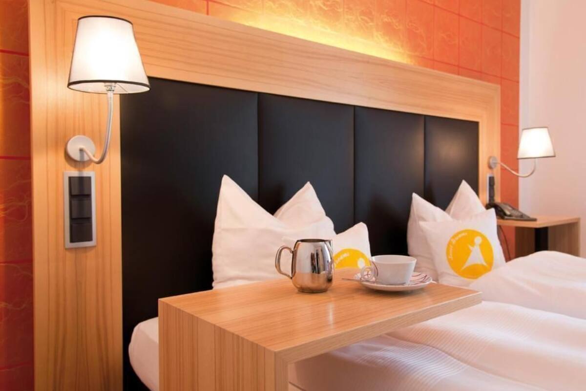 A hotel room with a bed, a bedside table with a lamp, a cup, and a mug.
