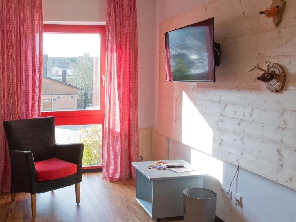 Habitació amb terra de fusta, una cadira negra amb coixí vermell, una taula petita amb un comandament a distància, televisió penjada a la paret i finestra amb cortines vermelles.