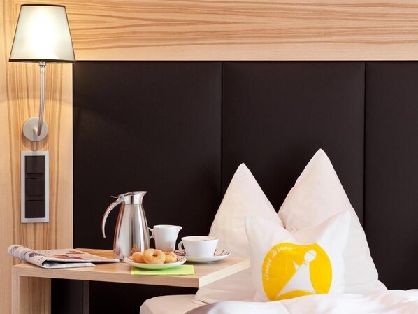 Una habitación de hotel con una cama, una mesita de noche con lámpara, una cafetera, tazas y pasteles.