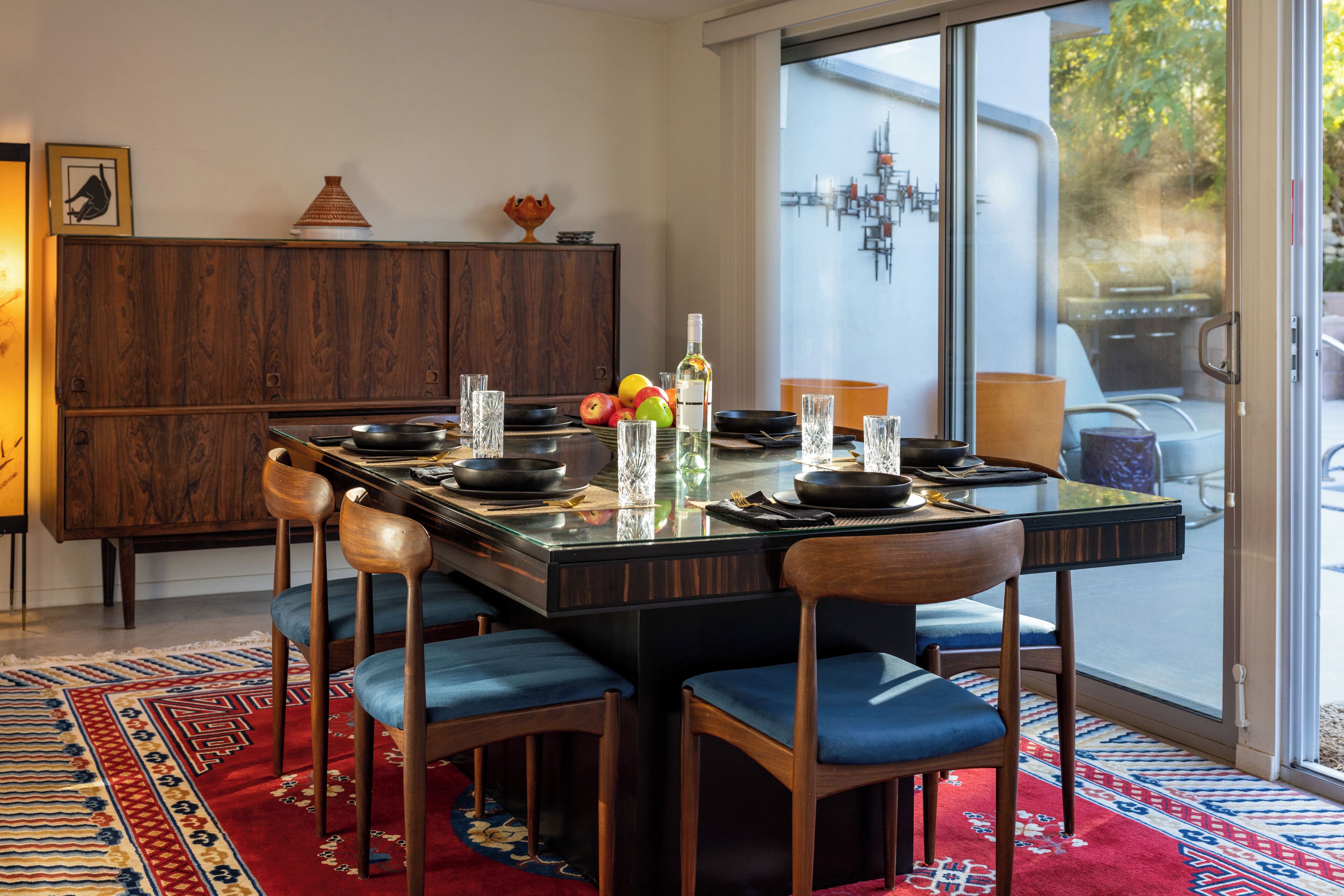 Een eetgedeelte met een glazen tafel, blauwe bekleden stoelen en een rood vloerkleed met patroon.