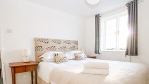 Een netjes opgemaakt bed met hoofdbord, nachtkastje met lamp, en raam met gordijnen.