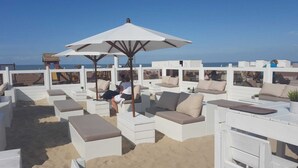 Zona relax in spiaggia con sedute imbottite bianche e un ampio ombrellone che offre ombra.