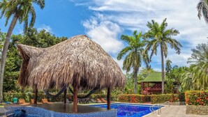A swimming pool with a thatched roof structure, surrounded by palm trees and a stone pathway. - Portales del Tayrona Garden Hotel (Santa Marta)