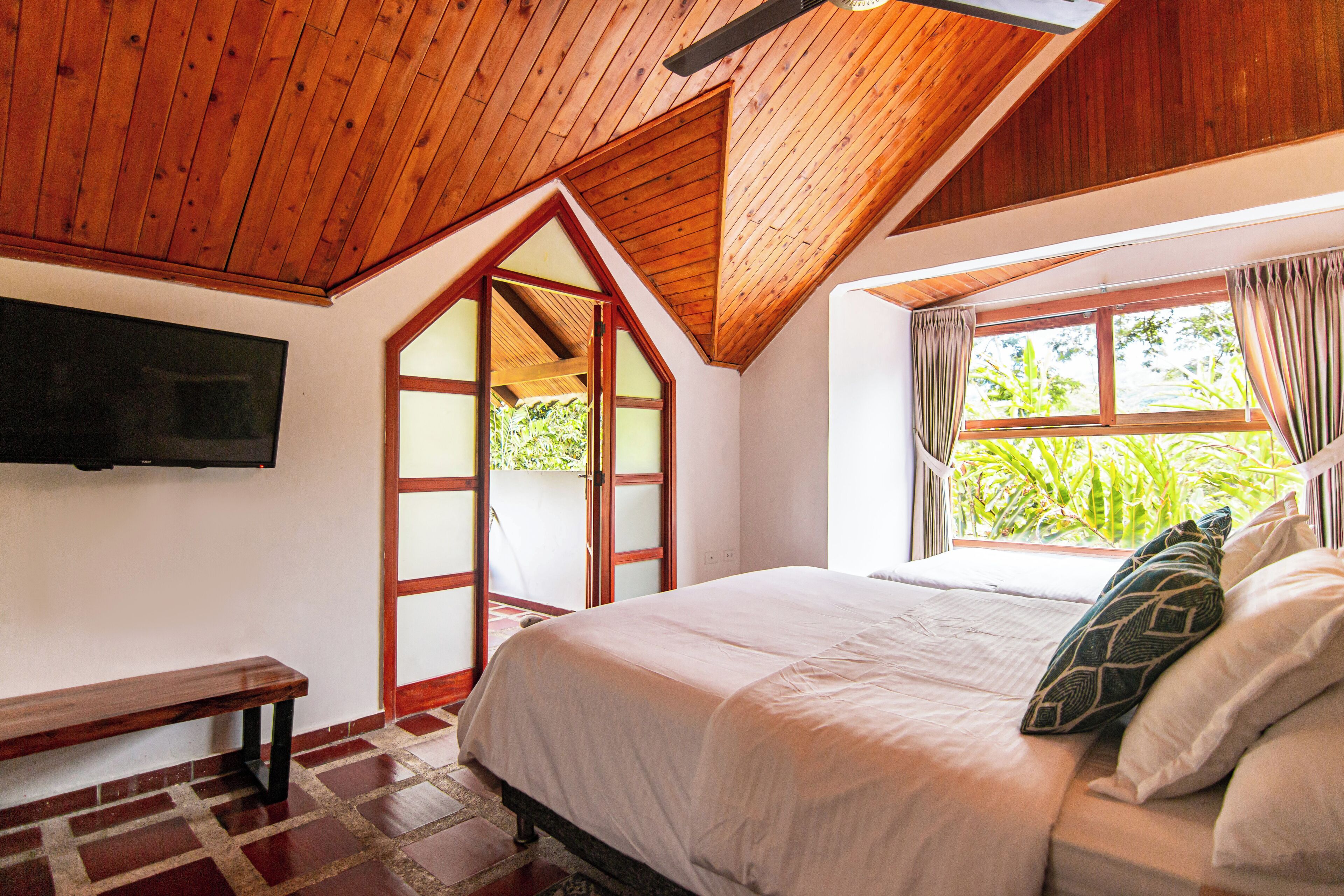 A bedroom with a wooden ceiling, a bed with white bedding, a bench, and a TV mounted on the wall.