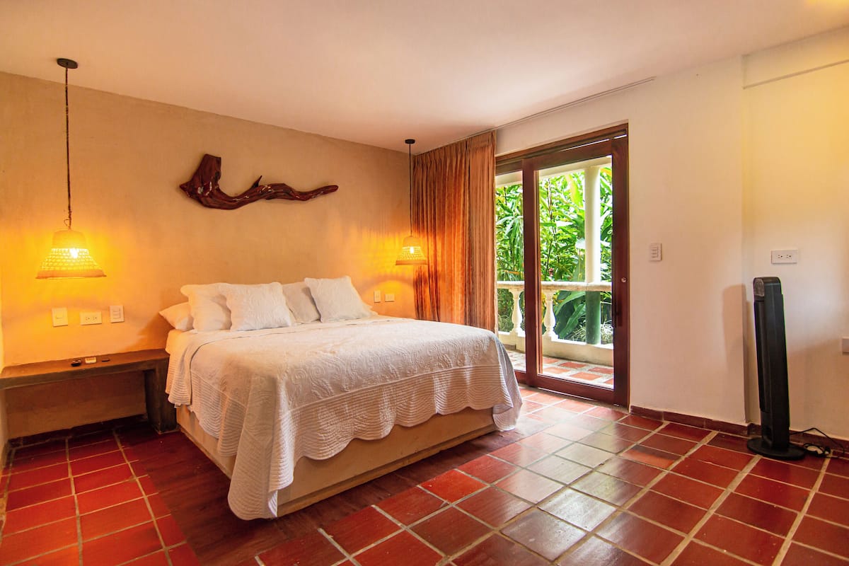 A bedroom with a bed, two bedside lamps, a bench, a wall-mounted decor piece, and a sliding glass door leading to a balcony.