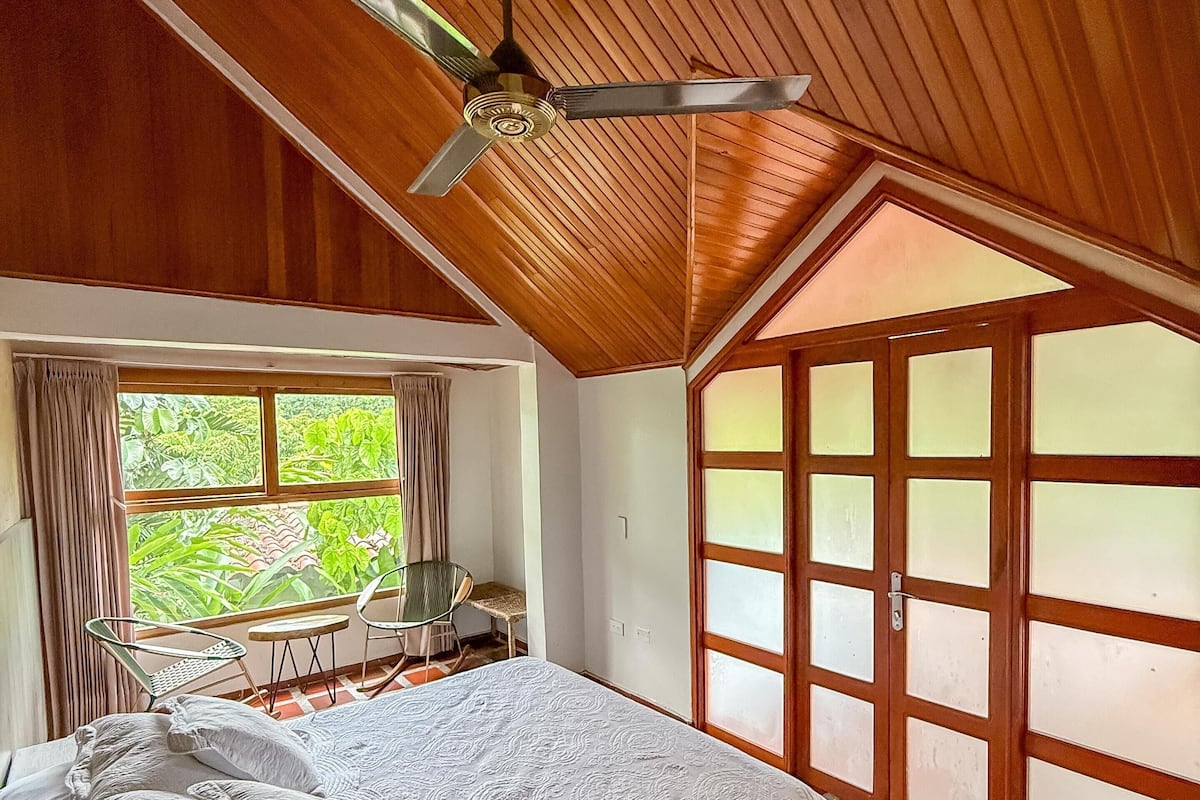 A bedroom with a wooden ceiling, a bed with a white bedspread, a ceiling fan, a window with curtains, and a view of greenery outside.