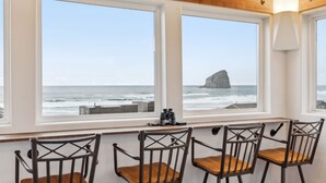 Una habitación con tres sillas de madera y una mesa, ventanas amplias con vistas al mar y una formación rocosa.