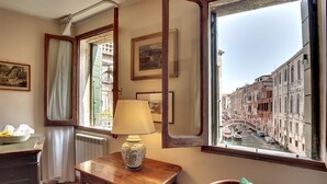 A room with a wooden desk, a lamp, a chair, a window with a view of a canal and buildings, and a radiator.