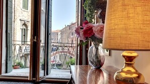 A room with a view of a canal and buildings, a wooden table with a lamp, and a vase with flowers.