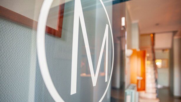 A close-up of a logo on a glass window, with a blurred interior view of a hallway and a staircase. - Hotel Monaco Zeebrugge Beach (Bruges)
