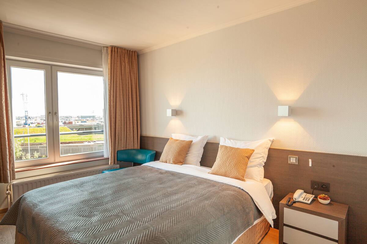 Une chambre d’hôtel avec un grand lit, une table de chevet avec un téléphone et un panier de fruits, une chaise bleue et une fenêtre avec des rideaux.