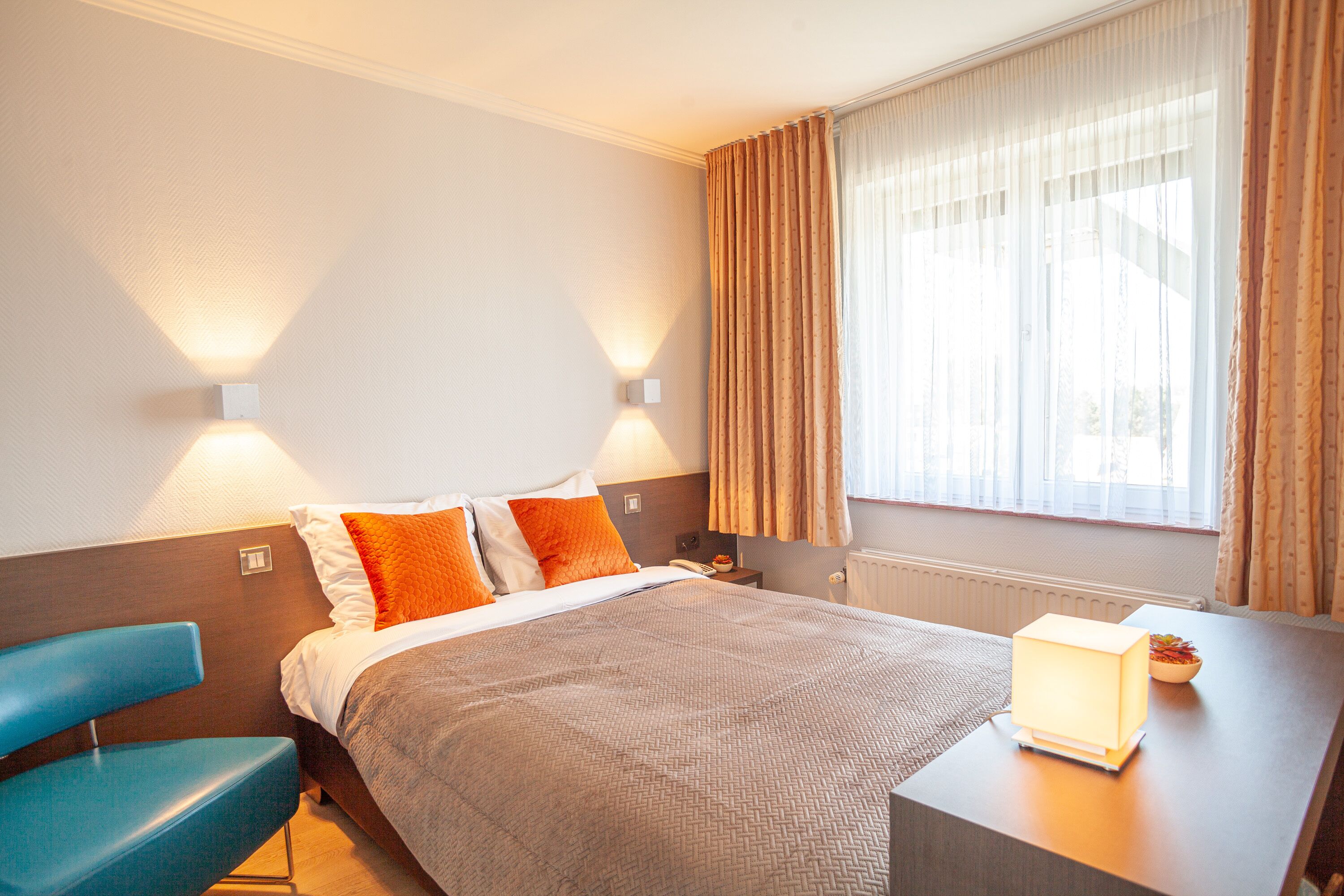 Quarto de hotel com cama, escrivaninha com luminária, cadeira e janela com cortinas.