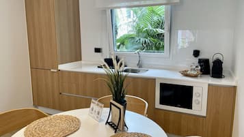 A modern kitchen with wooden cabinets, a white countertop, a sink, and a window with a view of greenery. There is a table with woven placemats and a vase with plants.
