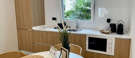 Una cocina moderna con armarios de madera, una encimera blanca, un lavamanos y una ventana con vistas a zonas verdes. Hay una mesa con individuales tejidos y un jarrón con plantas.