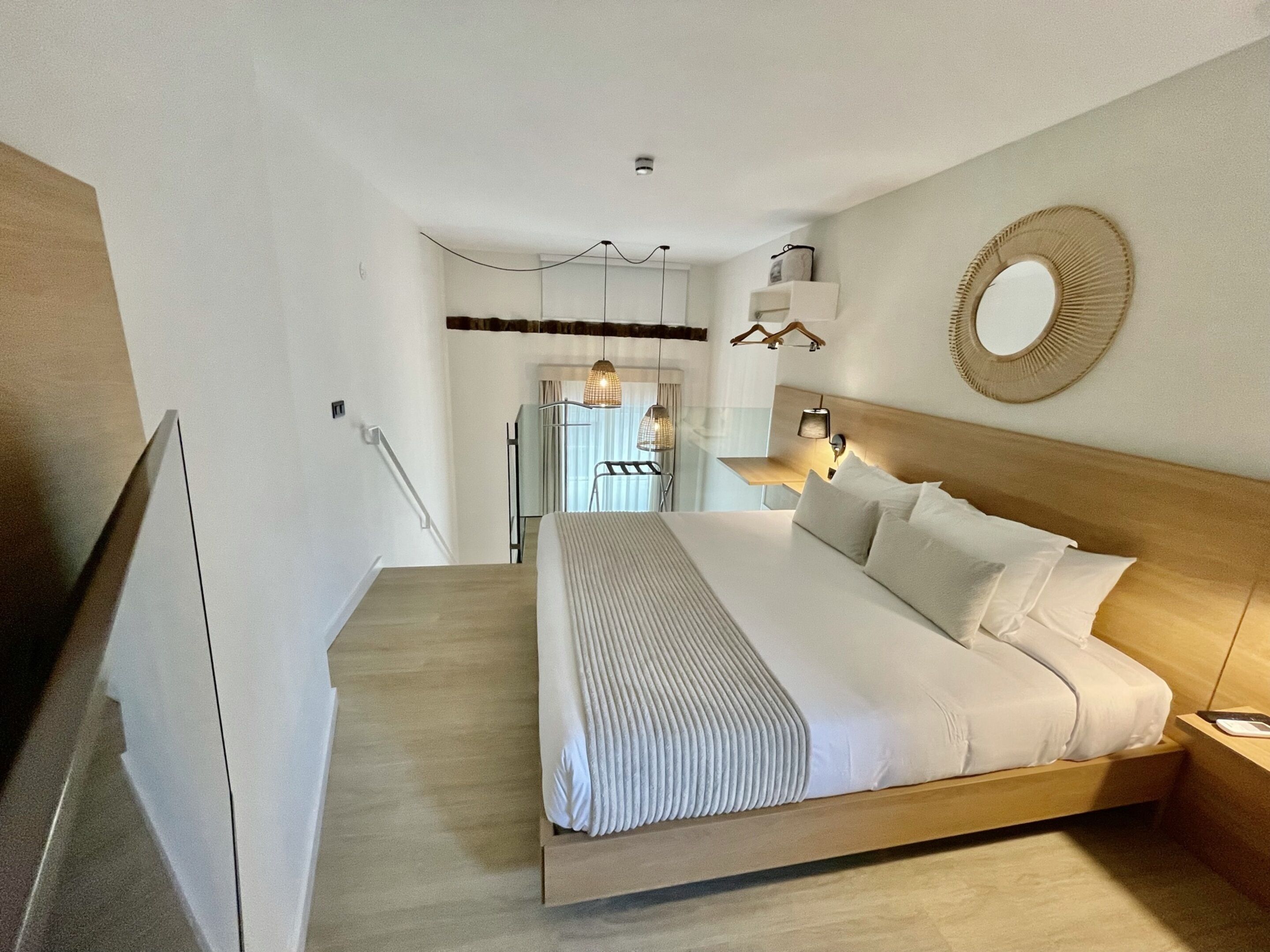 Une chambre d’hôtel moderne avec un grand lit, une tête de lit en bois et un grand miroir rond au mur.