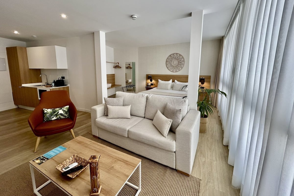 Una habitación de hotel moderna con sofá, una mesa de centro de madera, una silla roja, una cama con cabecero, una mesita de noche, una lámpara, un espejo y una ventana con cortinas.