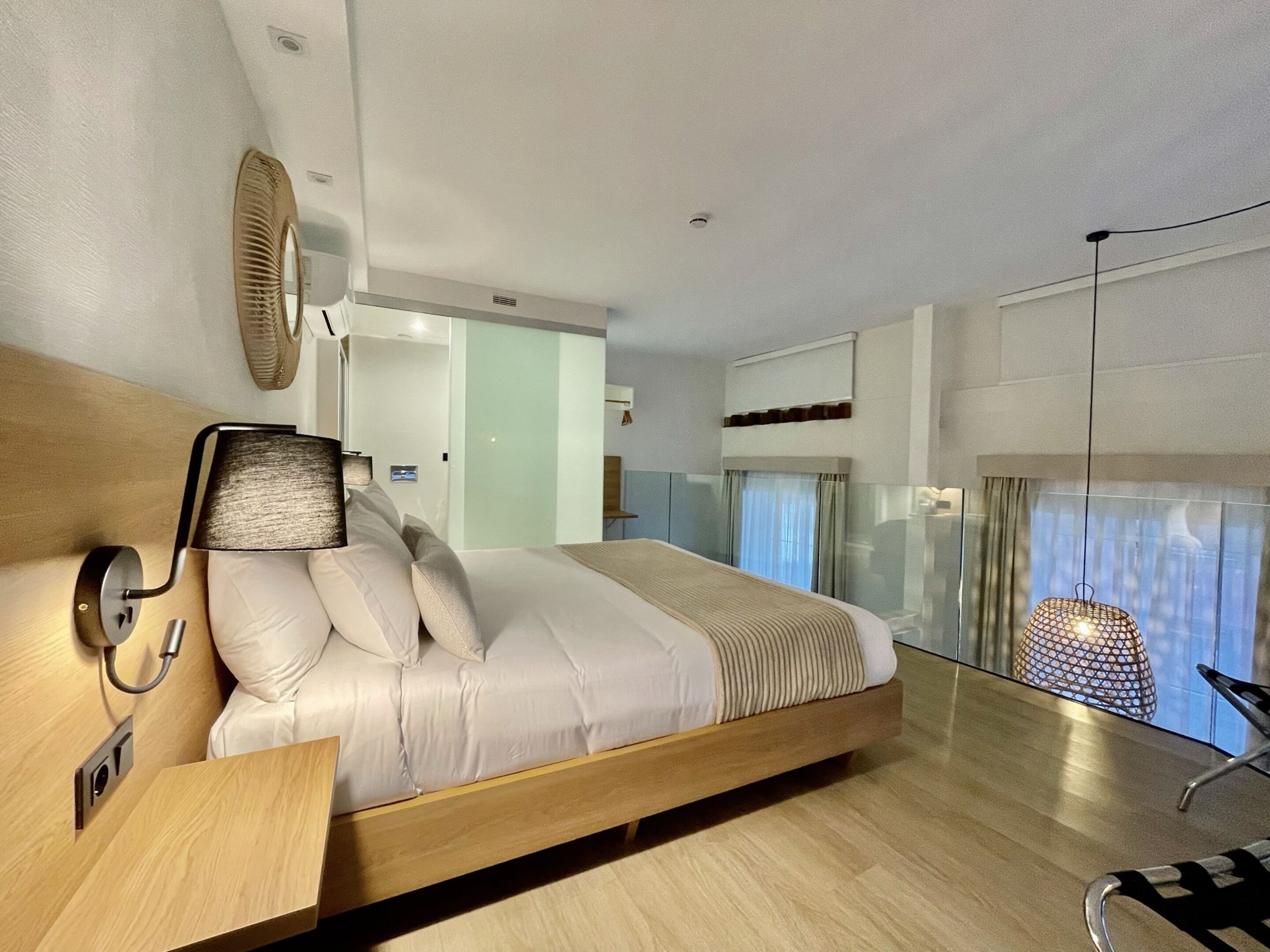 A modern bedroom with a wooden bed, a bedside table, a wall-mounted lamp, and a large window with a view.