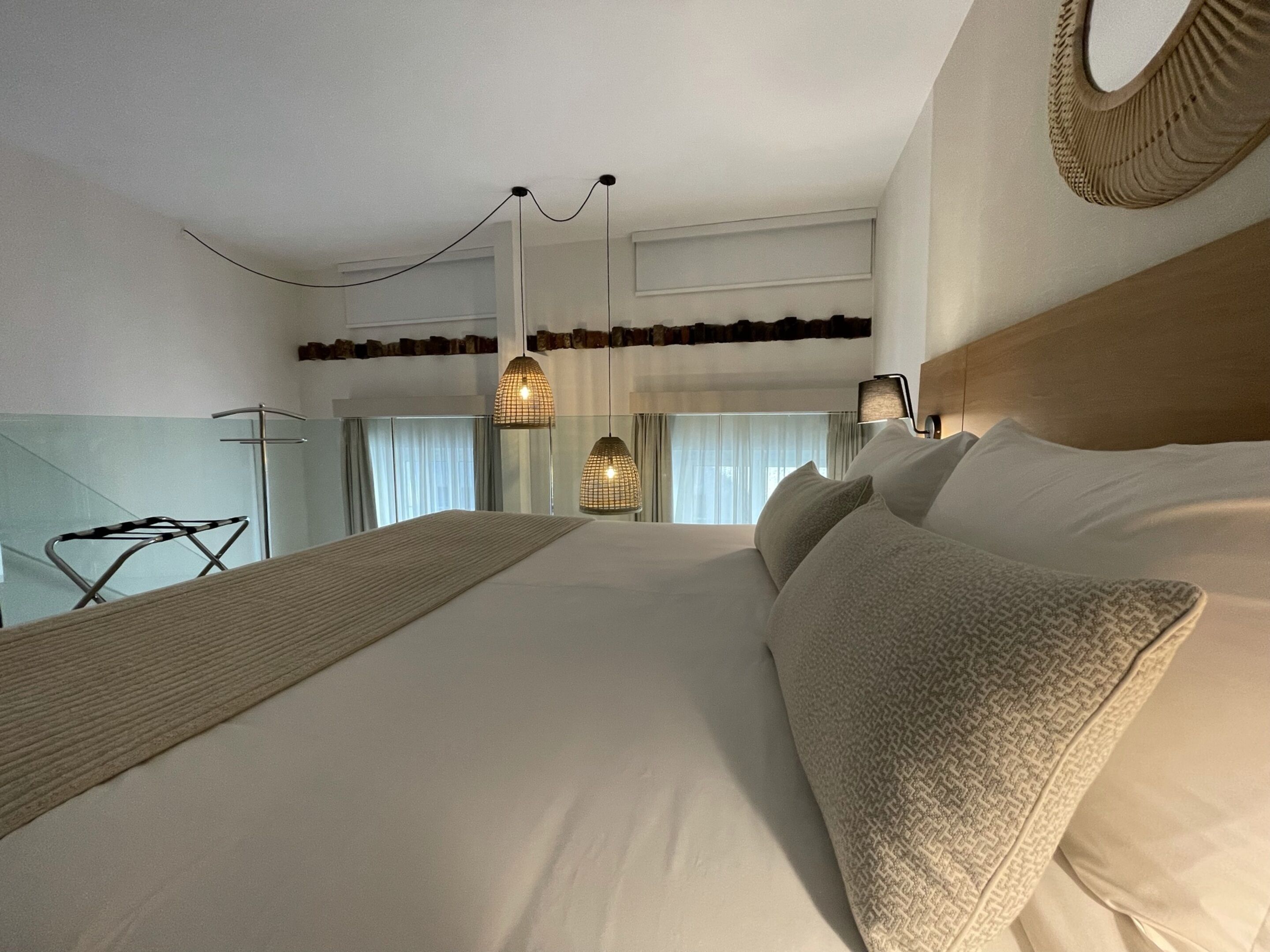 Habitación de hotel con una cama grande, cabecera de madera, lámpara colgante y vista a la ciudad por la ventana.