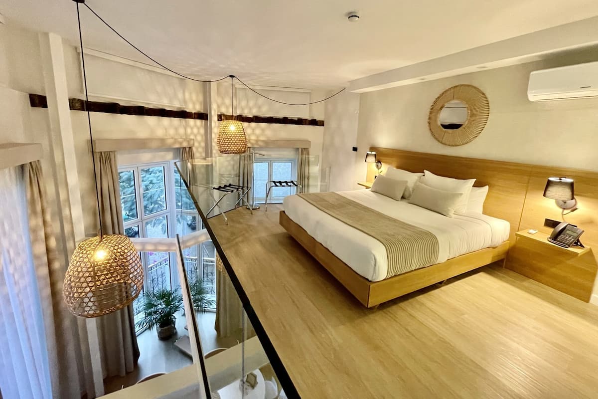 Habitación de hotel moderna con una cama grande, un separador de vidrio, un balcón con plantas y vistas al exterior.