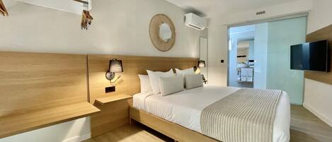 Une chambre d’hôtel moderne avec une tête de lit en bois, un grand lit avec une literie blanche, une table de chevet avec une lampe, un téléviseur fixé au mur et une petite étagère avec des objets décoratifs.