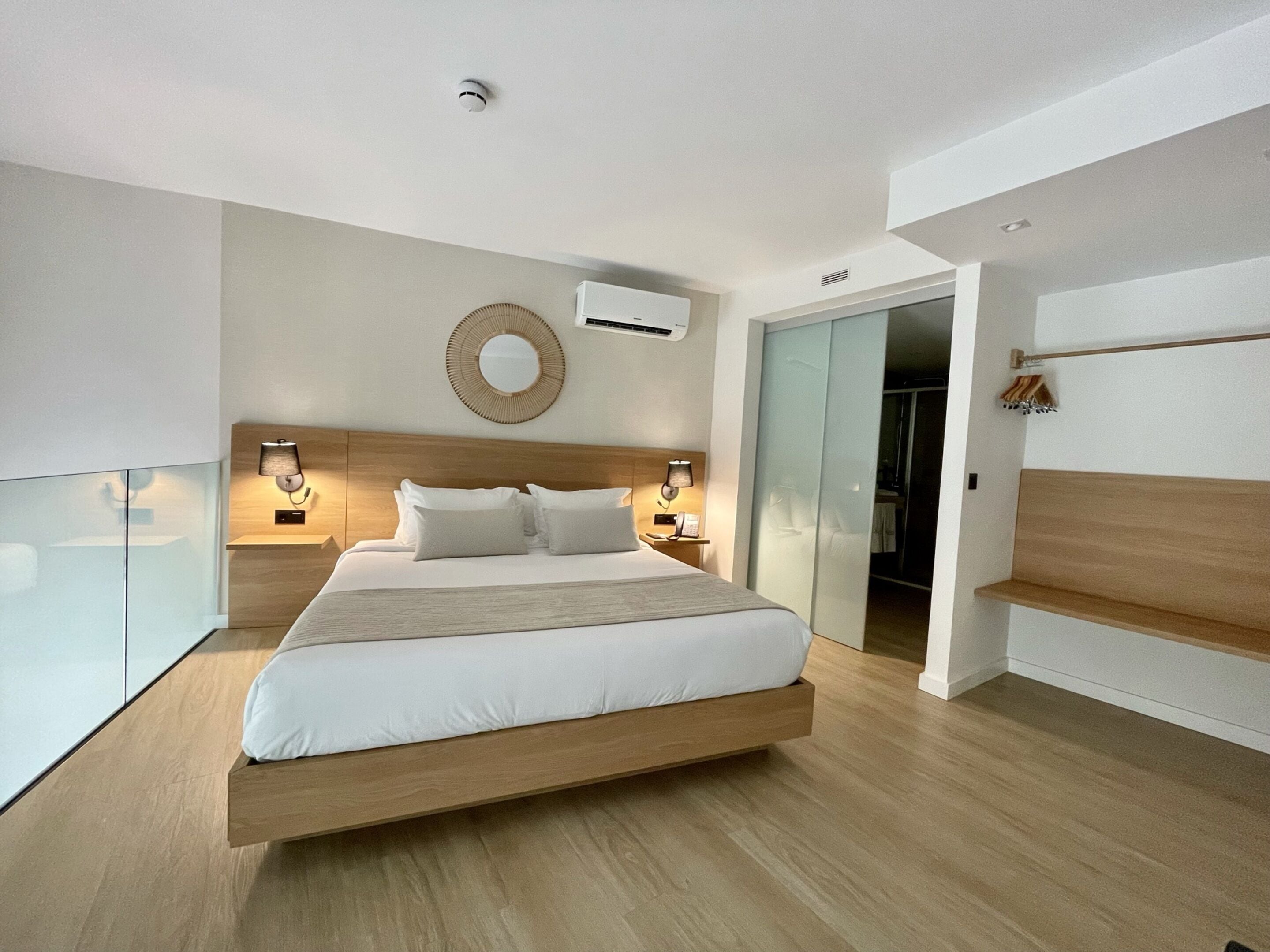 Une chambre d’hôtel moderne avec un grand lit, des lampes de chevet, un miroir rond et une cloison en verre.