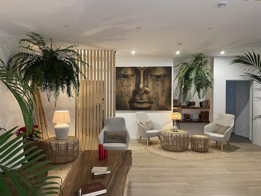 A modern living room with a wooden coffee table, two grey armchairs, a large abstract painting, and several potted plants.