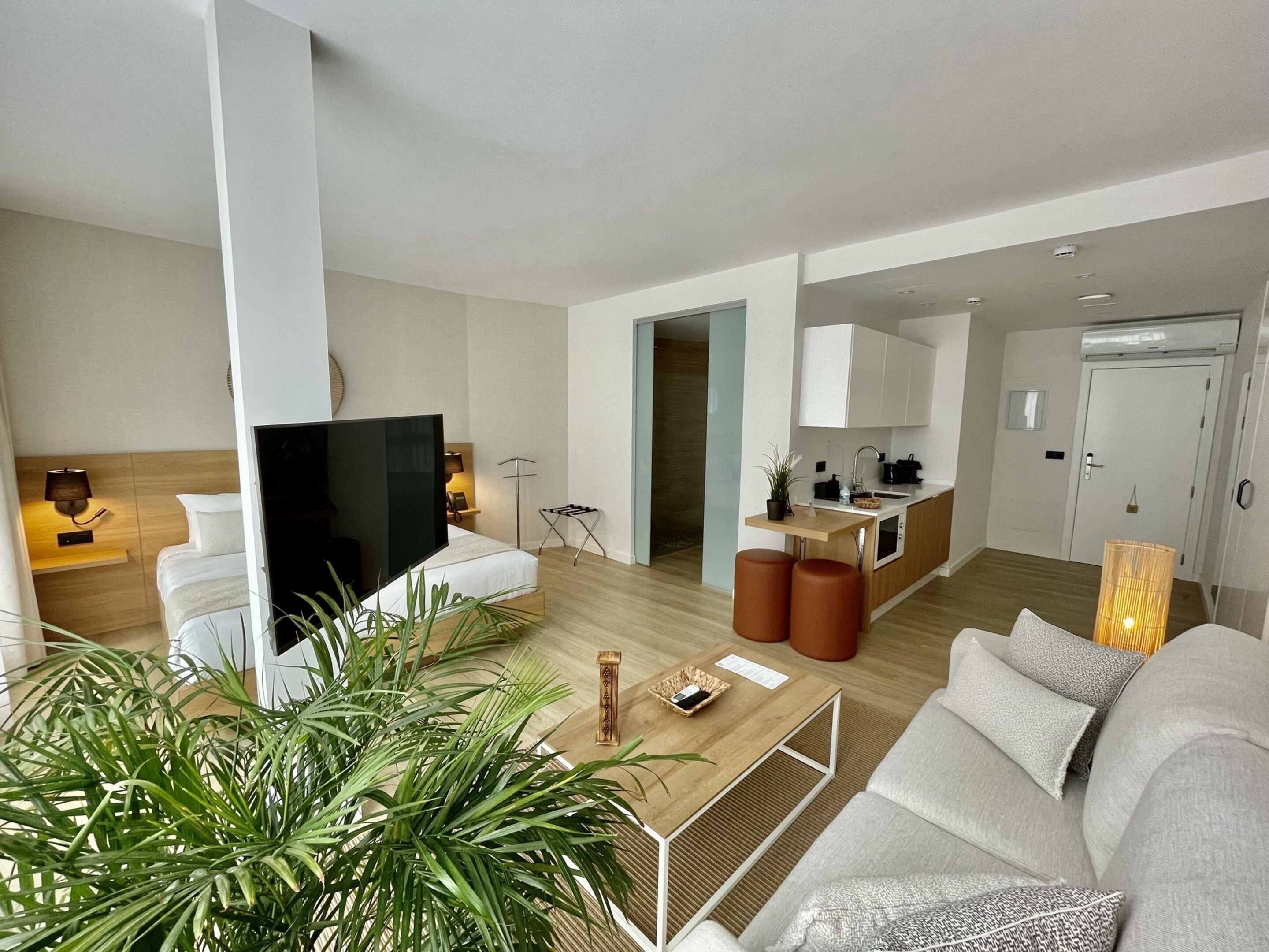 A modern living room with a sofa, a coffee table, and a TV. There is a kitchen area with cabinets and a sink.